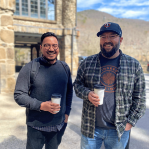 Amar and Nick standing outside with coffee cups in their hands smiling at the camera